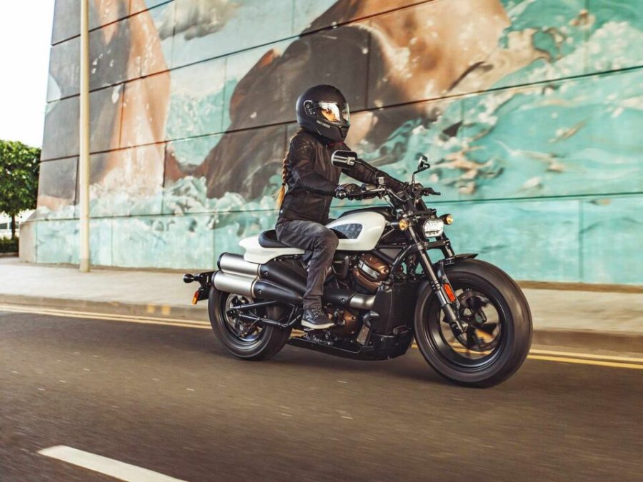 Una mujer con Harley-Davidson Sportster S blanca con llantas negras pasa por delante de un mural 