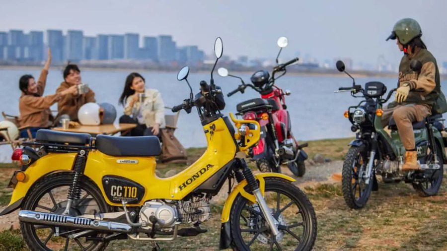 Una Honda Cross CUB 110 amarilla en primer plano y un grupo de jóvenes detrás, cerca de un lago. Una Honda Cross CUB 110 amarilla en primer plano y un grupo de jóvenes detrás, cerca de un lago.