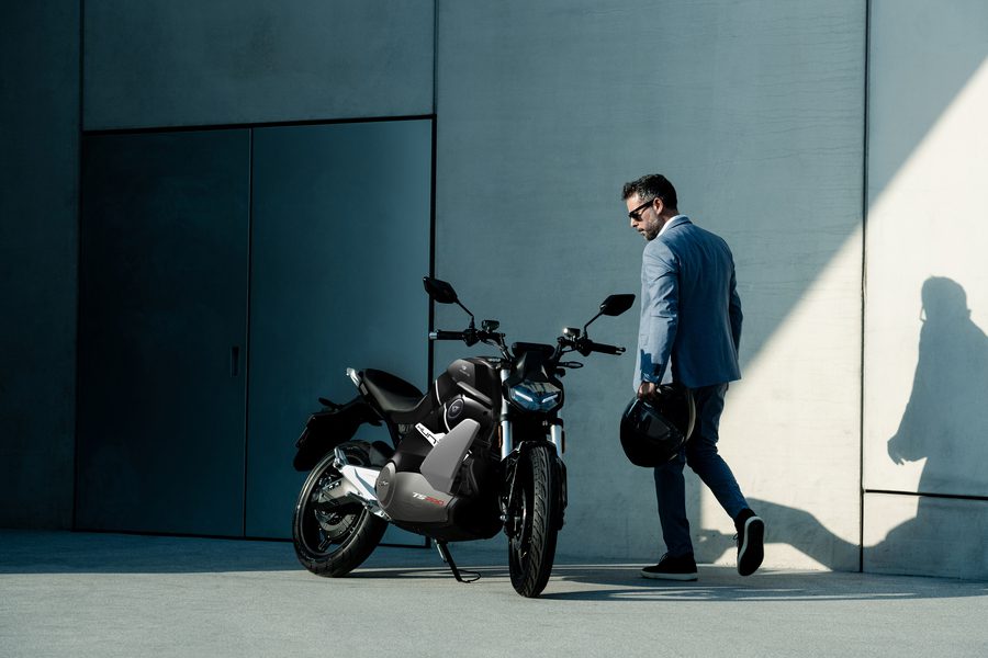 Un joven de saco celeste, jeans y zapatillas se acerca a una VMoto Hunter negra y gris