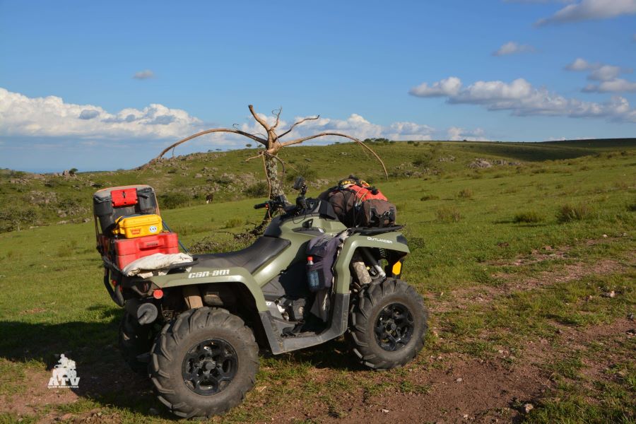 Un ATV verde con equipaje en medio de un terreno montañoso pero con pastos naturales.