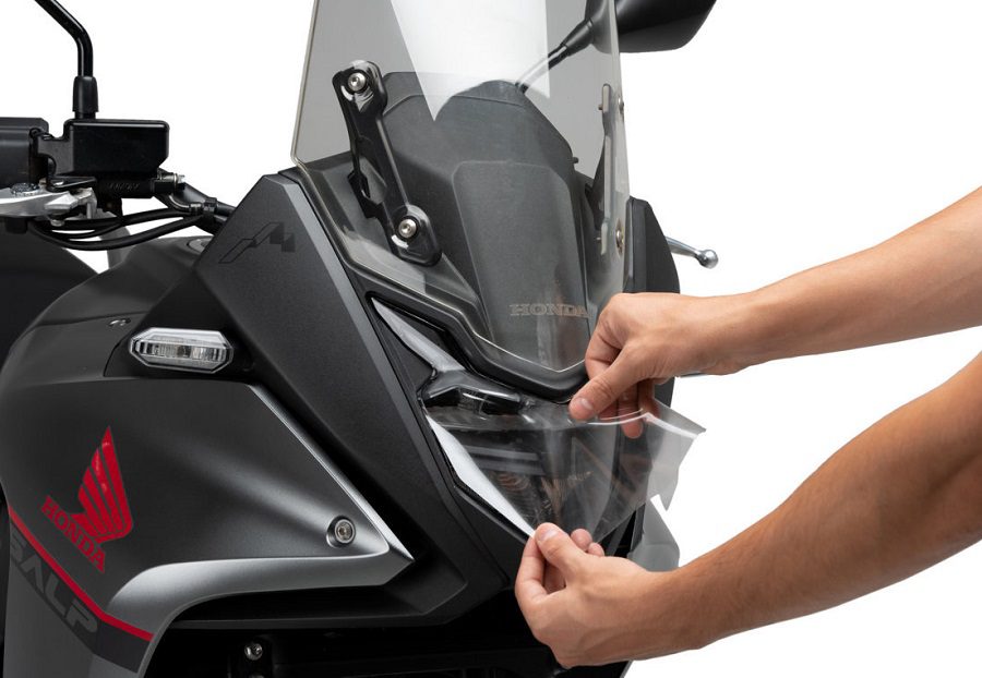 Una persona aplica la lámina del protector de faro Adhere a una Honda Transalp gris.