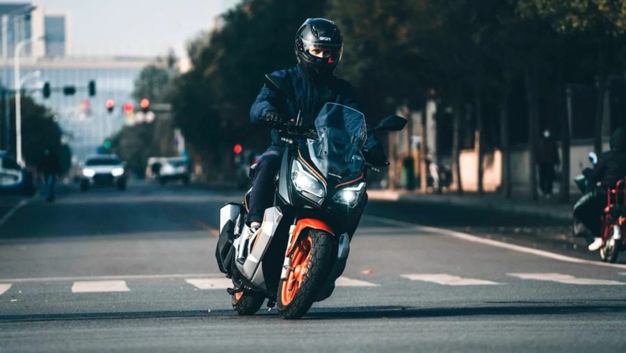 Un scooter QJ Motor ATR 125 gris y naranja avanza de frente por una calle. Un scooter QJ Motor ATR 125 gris y naranja avanza de frente por una calle.
