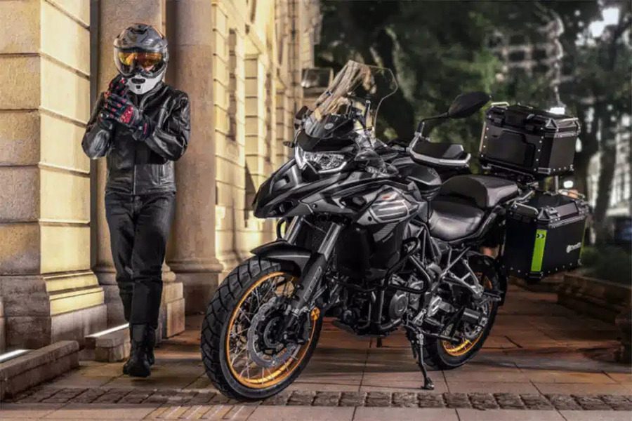 Un piloto se acomoda el casco al lado de una Benelli TRK 552 gris y llantas doradas.