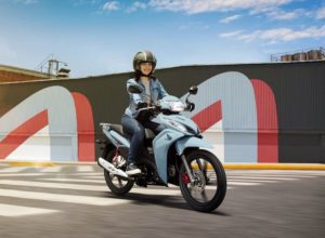 Una mujer se deplaza en una Honda Wave celeste con fondo de pared con graffitti