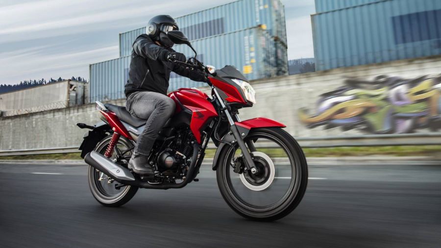 Un motociclista con chaqueta negra y pantalones de jeans grises avanza por la ciudad en una Honda CB125F Twister roja