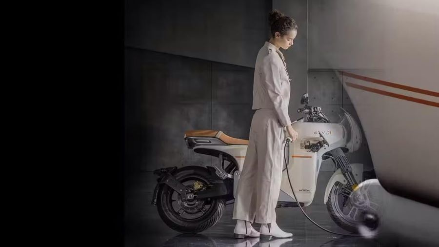 Una jovencita vestida de blanco y a tono con la Café Racer Honda E-VO, simula introducirle el cable de carga