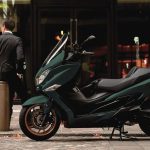 El scooter Suzuki Burgman 400 siempre se renueva Un scooter Suzuki Burgman 400 modelo 2025 de color verde intenso permanece estacionado en la calle mientras su dueño, de riguroso traje oscuro, descansa en segundo plano