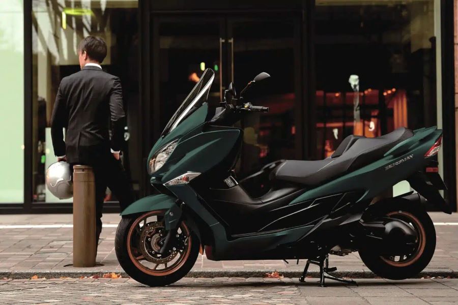 Suzuki-Burgman 400 2025 Un scooter Suzuki Burgman 400 modelo 2025 de color verde intenso permanece estacionado en la calle mientras su dueño, de riguroso traje oscuro, descansa en segundo plano