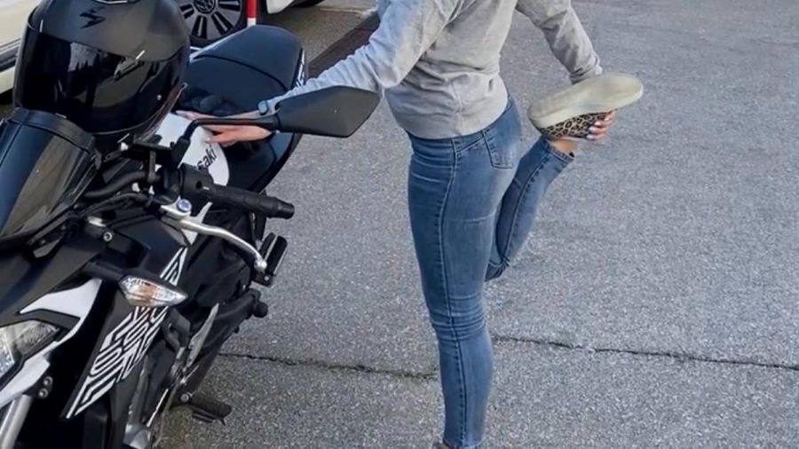 Una mujer con jeans y remera gris elonga su pierna derecha antes de subirse a la moto
