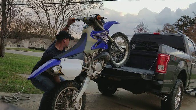 subiendo moto dr crros a camioneta Un hombre intentando subir solo una moto de cross a la caja de una camioneta.