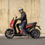 Un usuario con vestimenta sport y casco jet se apresta a salir con un scooter eléctrico naranja