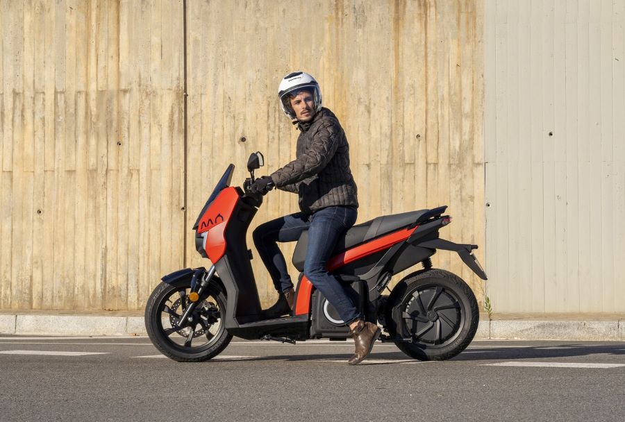 Un usuario con vestimenta sport y casco jet se apresta a salir con un scooter eléctrico naranja