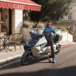 Una mujer joven chequea su celular sentada sobre un BMW CE 04 y frente a un bar