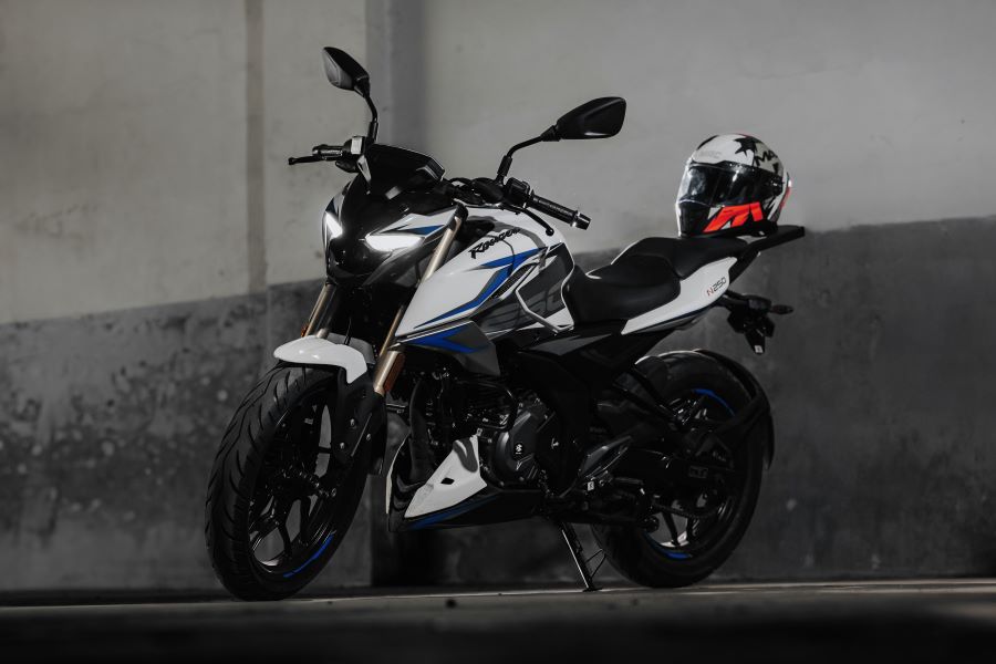 Una Bajaj Rouser N250 UG 2025 de color blanco con vivos azul estacionada y con un casco sobre el asiento