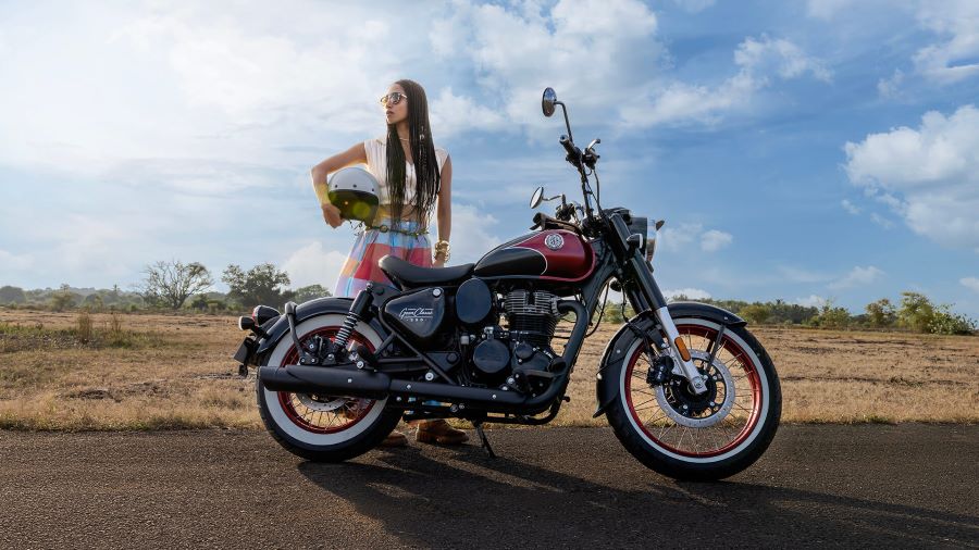 Una mujer con el casco en la mano mira hacia el infinito detrás de una Royal Enfield Goan Classic 350 roja y negra