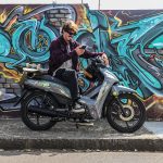 Un joven consulta su celular sentado sobre una Zanella DUE ST gris y con una pared con grafiti de fondo