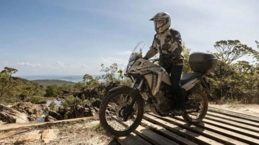 Un piloto experimentado se para sobre los reposapies de una Honda Sahara XRE 300 blanca para sortear las tablas de un puente rústico