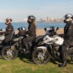 La Corven Triax 250 Adventure viene lista para viajar Tres usuarios, parados en forma paralela sobre sus Corven Triax 250 Adventure, observan el horizonte con el mar y la ciudad de fondo