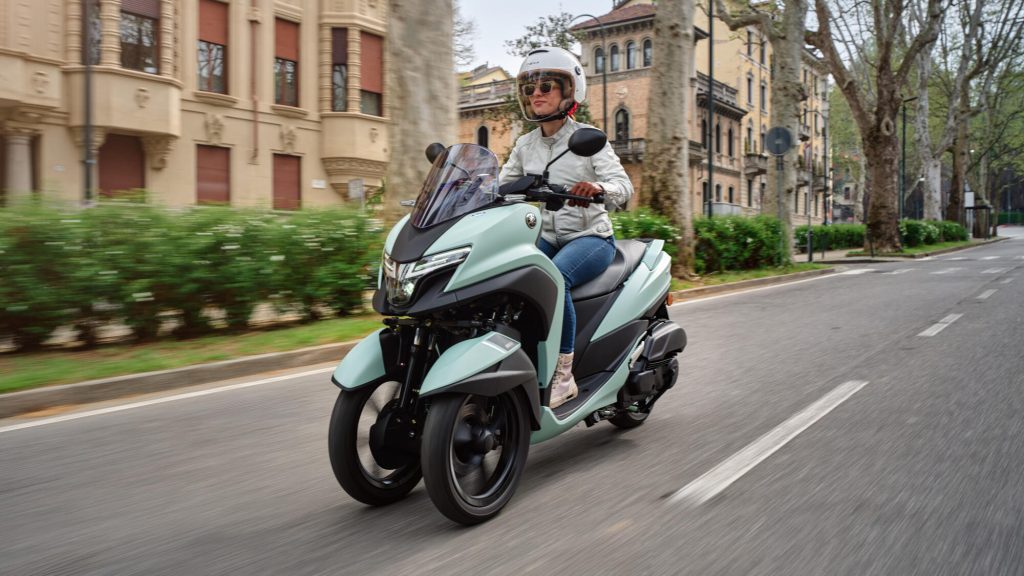 Una mujer joven conduce una Yamaha Tricity 125 verde en un sector urbano con edificios históricos