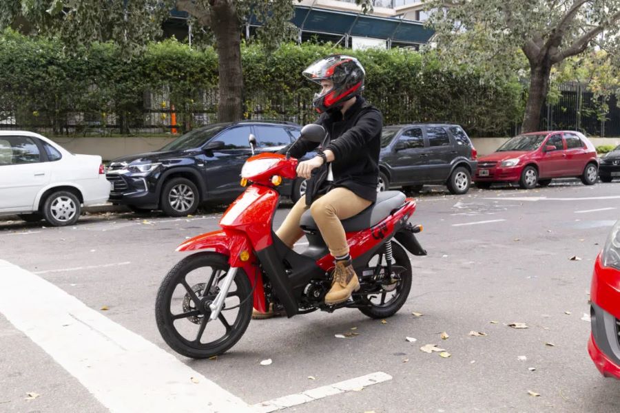 Un piloto joven con ropa informal espera la apertura del semáforo sobre una moto CUB 110 roja