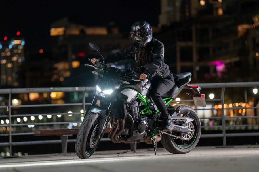 Una mujer sentada sobre una Kawasaki Z900 2025 de noche, con las luces encendidas y una baranda de fondo