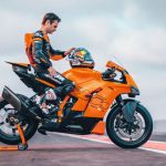 KTM 990 RC R: poder sin excesos en modo supersport Dani Pedrosa se concentra con la vista hacia abajo y su mano izquierda sobre el casco, sentado sobre una KTM 990 RC R naranja dispuesta en la calle de un paddok