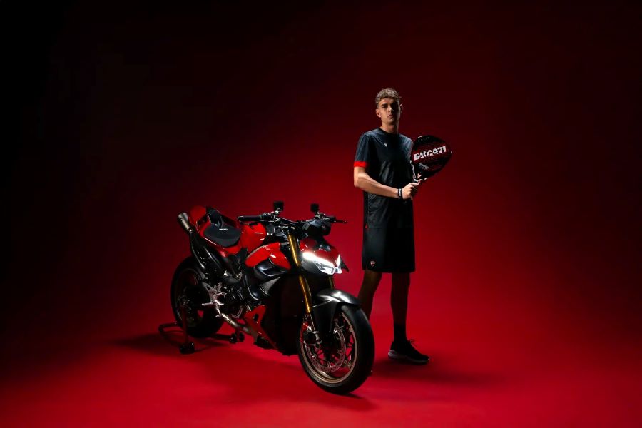 Un modelo posa con indumentaria MaceonXDucati negra y roja al lado de una superdeportiva de la marca