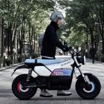 Un usuario joven cruza verticalmente una calle llevando de la mano un prototipo de scooter Suzuki e-vanvan