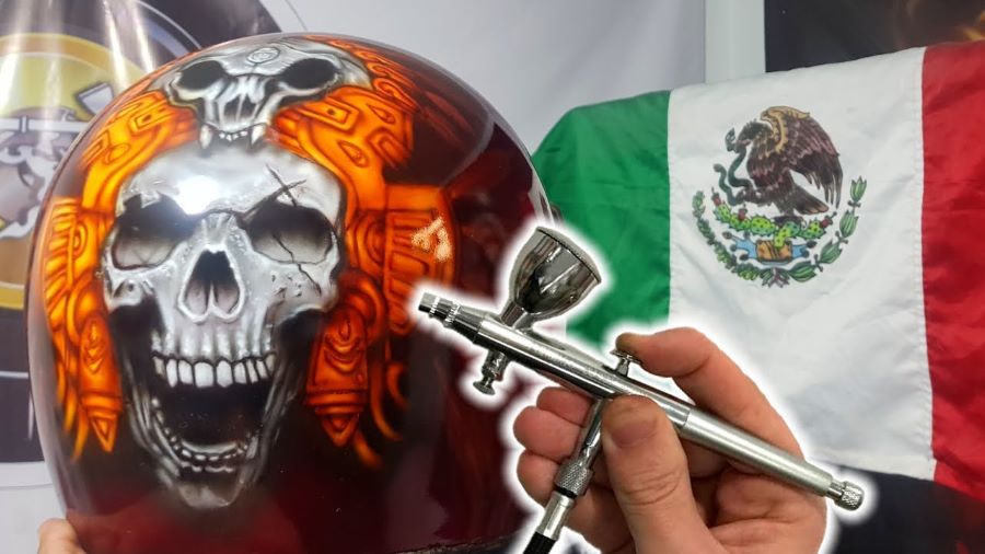 Imagen donde se ven un aerógrafo y un casco intervenido con una figura de calavera con la bandera de México de fondo