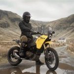 Un piloto con ropa oscura posa sobre una moto BSA Thunderbolt amarilla en un entorno montañoso