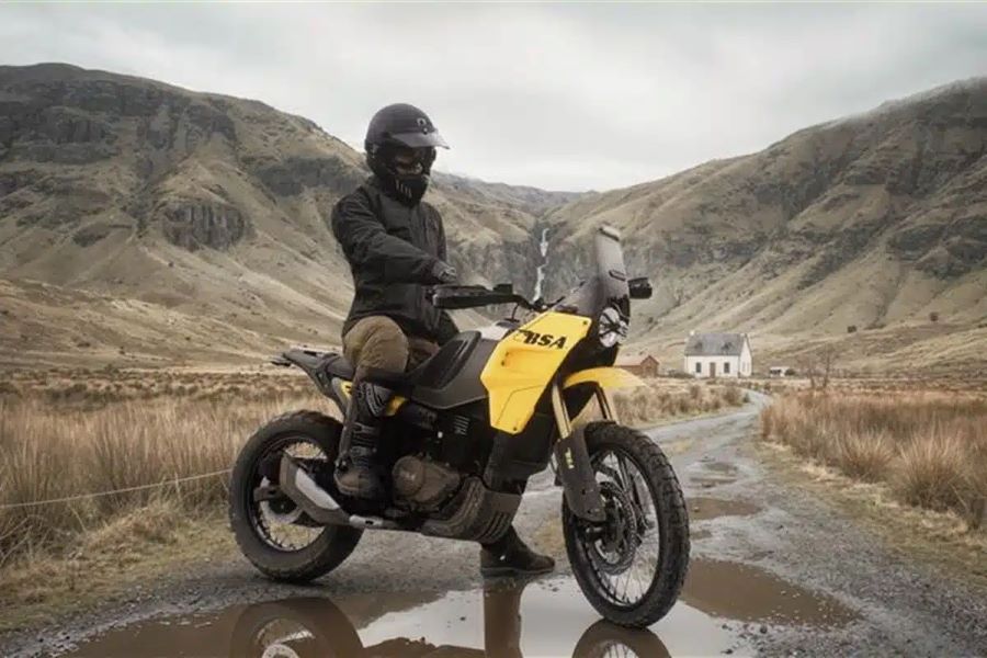 Un piloto con ropa oscura posa sobre una moto BSA Thunderbolt amarilla en un entorno montañoso