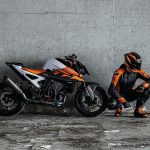 Un piloto posa en cuclillas al lado de una TM 990 Duke 2026 con una pared gris de fondo