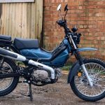 Vista lateral derecha de una moto AJS Imber 125 azul con fondo de pared de ladrillos