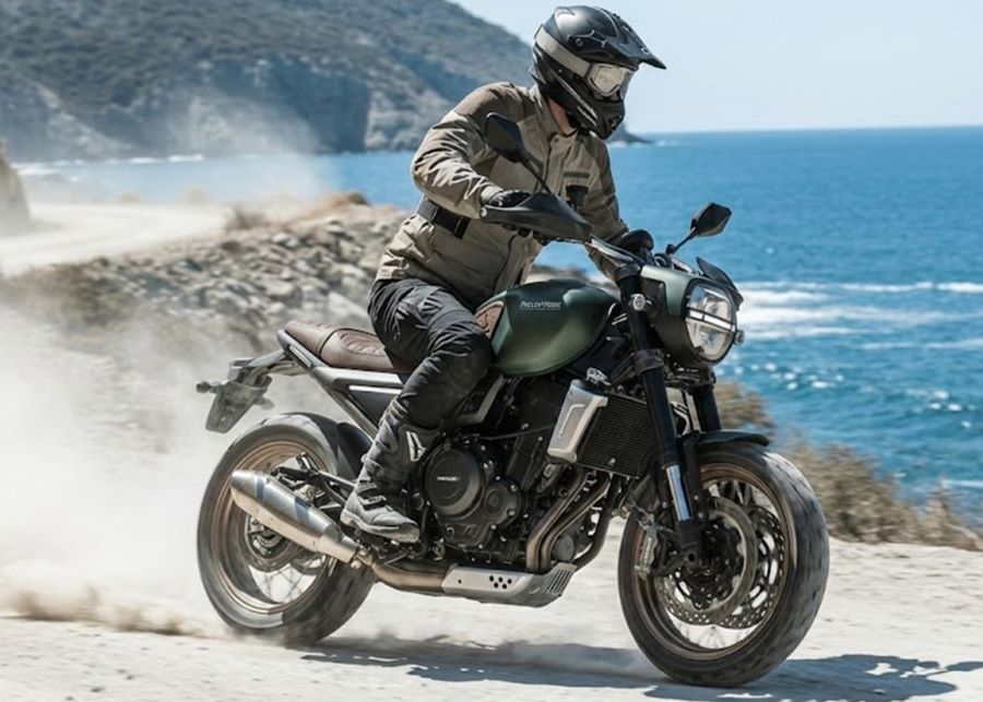 Un piloto toma un curva de una ruta de tierra bordeada por el mar sobre una moto Phelon & Moore Brighton 6 scrambler