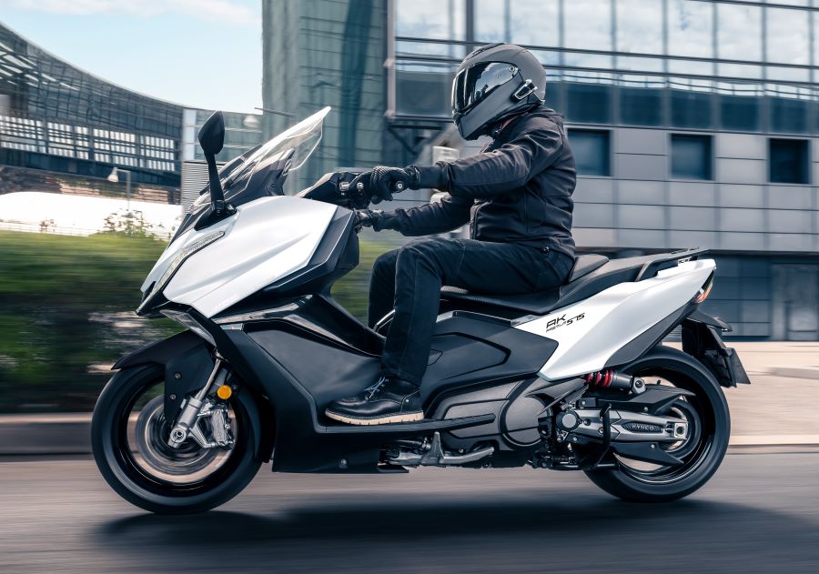 Un scooter Kymco AK Premiun 575 gris metalizado circula por una calle urbana