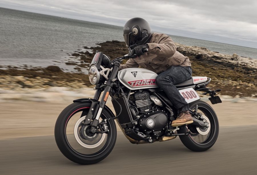 Una moto Triumph Tracker 400 blanca se desplaza por una ruta que bordea un espejo de agua
