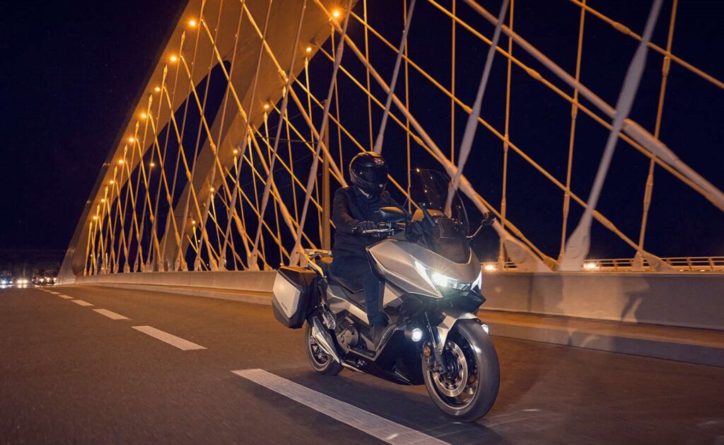 Un scooter Honda Forza 750 de desplaza de noche, con la luces encendidas, sobre un puente carretero