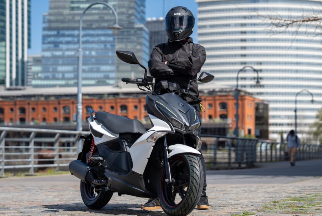 Un usuario, vestido de negro y con los brazos cruzados, aguarda detrás de una scooter Kymco Super 8 150 blanco al frente de edificios modernos