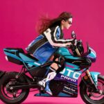 En una foto de estudio, una joven piloto, con ropa de calles, simula viajar a toda velocidad sobre una CFMoto XO Papio Racer 2026 azul