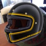 Un casco de moto negro intervenido con una línea amarilla pintada que recorre el exterior de visera y la base del mismo.
