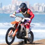 Un pìloto de competición surca la arena con el mar y una ciudad de fondo