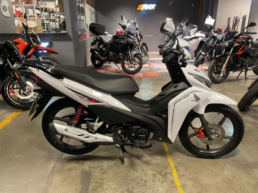 En primer plano, una moto Honda Wave 110 blanca dentro de un local donde aparecen más unidades de otro carácter
