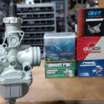 Una mano muestra en primer plano un carburador de moto con fondo de cajas de distiintas marcas