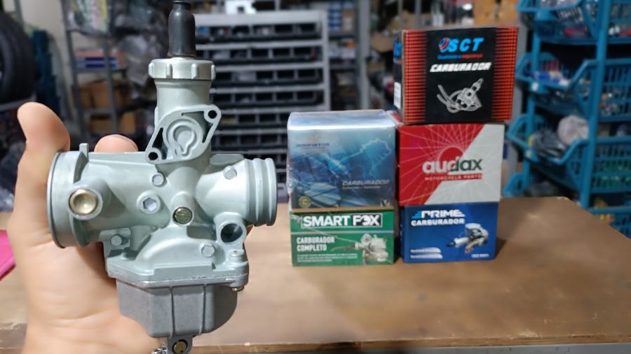 Una mano muestra en primer plano un carburador de moto con fondo de cajas de distiintas marcas