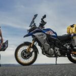 Charly Sinewan ¿Por qué es una referencia de viajes en moto? charly sinewan, de espaldas, posa junto a su moto ante un paisaje montañoso
