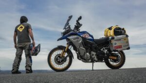 charly sinewan, de espaldas, posa junto a su moto ante un paisaje montañoso