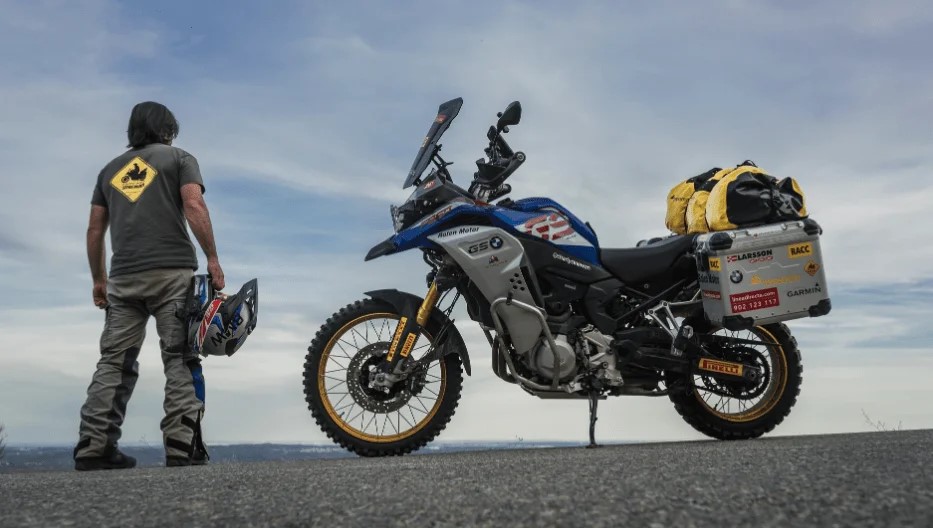 charly sinewan, de espaldas, posa junto a su moto ante un paisaje montañoso