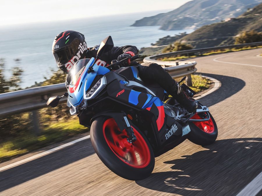 una Aprilia RS 457 azul con llantas naranja toma una curva en una carretera