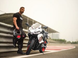 Un piloto, casco en mano, se apoya en el guardrail al lado de su moto Aprilia RS 457 blanca