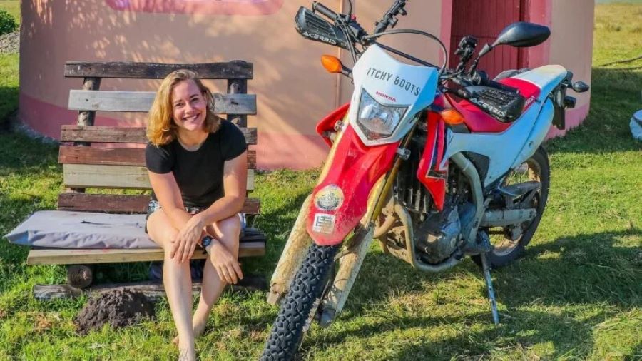 Itchy Boots sentada en un banco de madera al lado de una moto offroad roja